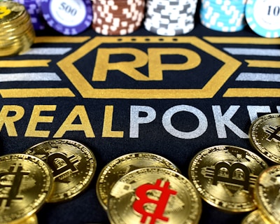 Stacks of poker chips in various colors are arranged in the background, while in the foreground, there are numerous golden coins featuring the Bitcoin symbol. The text 'REALPOKER' is prominently displayed in gold and silver lettering on a dark background.