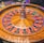 A roulette wheel with alternating red and black numbered pockets on a wooden platform. The wheel contains a small white ball resting in one of the pockets, indicating gameplay. Surrounding the wheel are stacks of colorful poker chips, suggesting a casino environment.