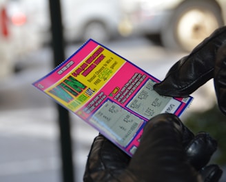 A person wearing black gloves is holding a colorful lottery scratch-off ticket. The ticket includes information about a $1,000,000 guaranteed minimum payout and features multiple games with various prize amounts.